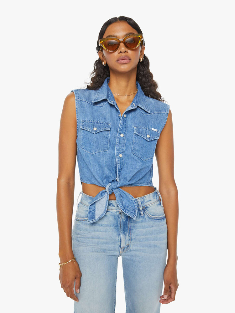 Front view of a woman medium blue sleeveless button-up with front patch pockets and a cropped hem that ties.