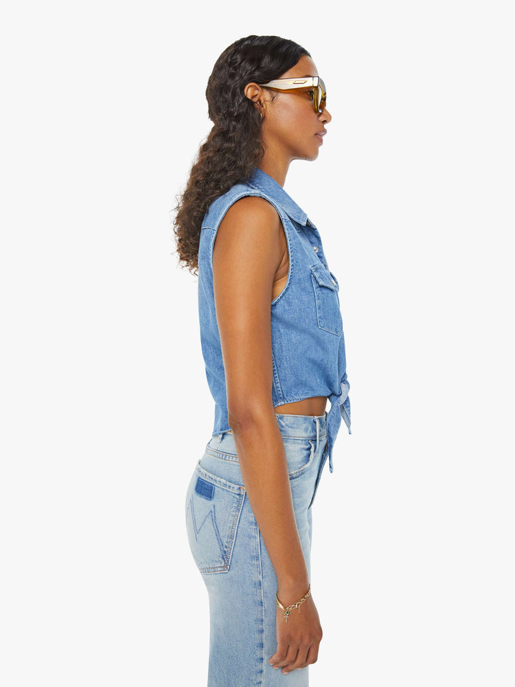 Side view of a woman medium blue sleeveless button-up with front patch pockets and a cropped hem that ties.