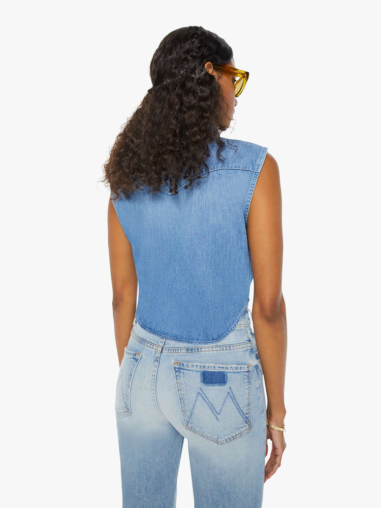 Back view of a woman medium blue sleeveless button-up with front patch pockets and a cropped hem that ties.