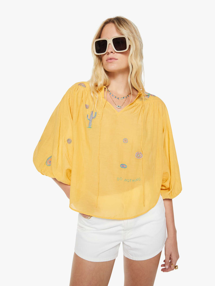 Front view of a woman in a golden yellow with colorful embroidery lightweight blouse with a V-neck that ties, 3/4-length balloon sleeves, a curved hem and ruffles throughout for a loose, flowy fit.