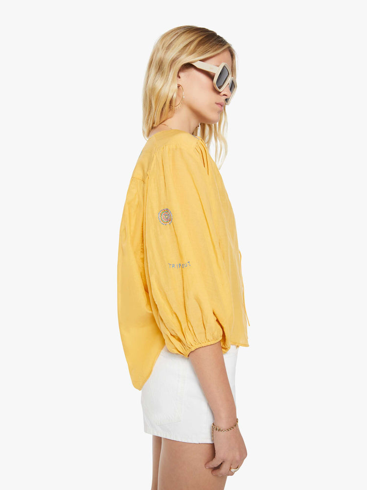 Side view of a woman in a golden yellow with colorful embroidery lightweight blouse with a V-neck that ties, 3/4-length balloon sleeves, a curved hem and ruffles throughout for a loose, flowy fit.