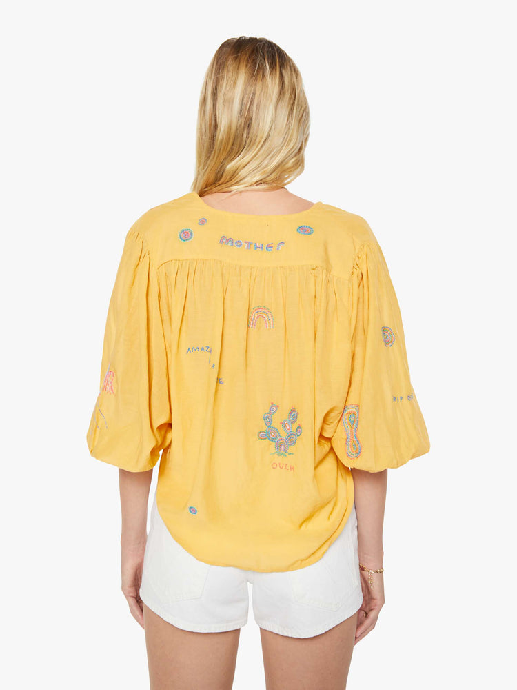 Back view of a woman in a golden yellow with colorful embroidery lightweight blouse with a V-neck that ties, 3/4-length balloon sleeves, a curved hem and ruffles throughout for a loose, flowy fit.
