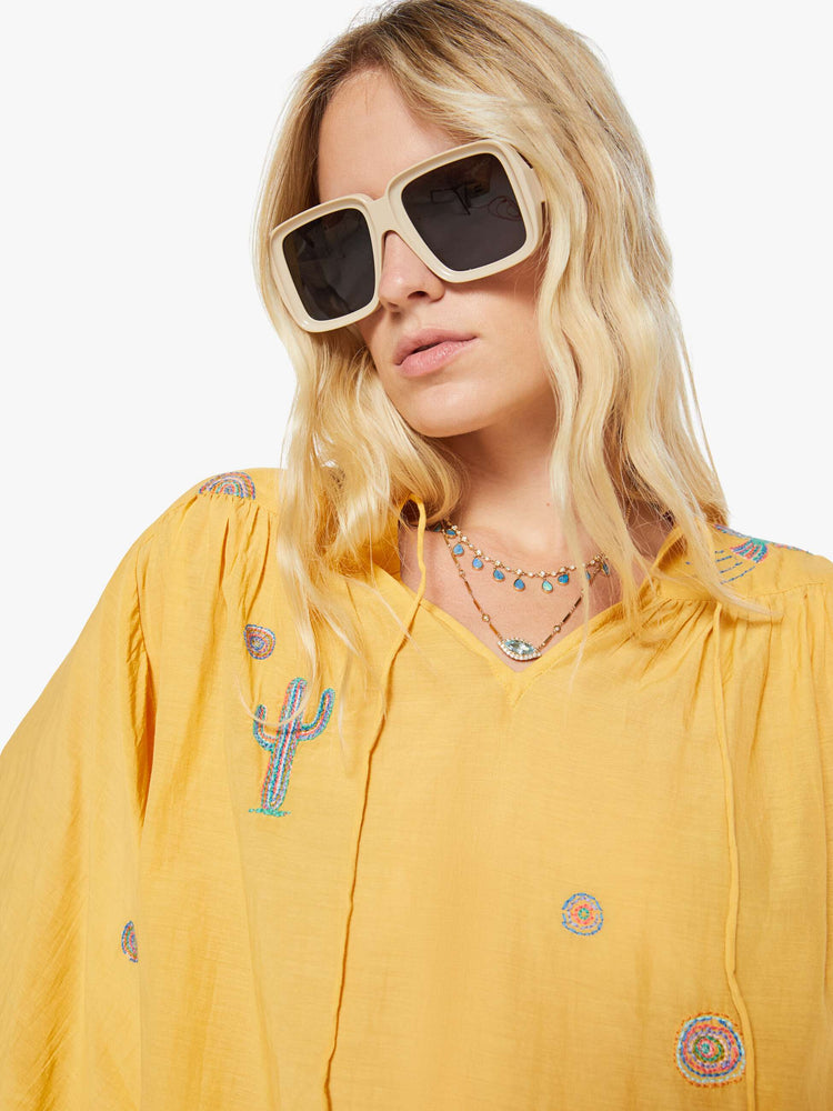 Close up view of a woman in a golden yellow with colorful embroidery lightweight blouse with a V-neck that ties, 3/4-length balloon sleeves, a curved hem and ruffles throughout for a loose, flowy fit.