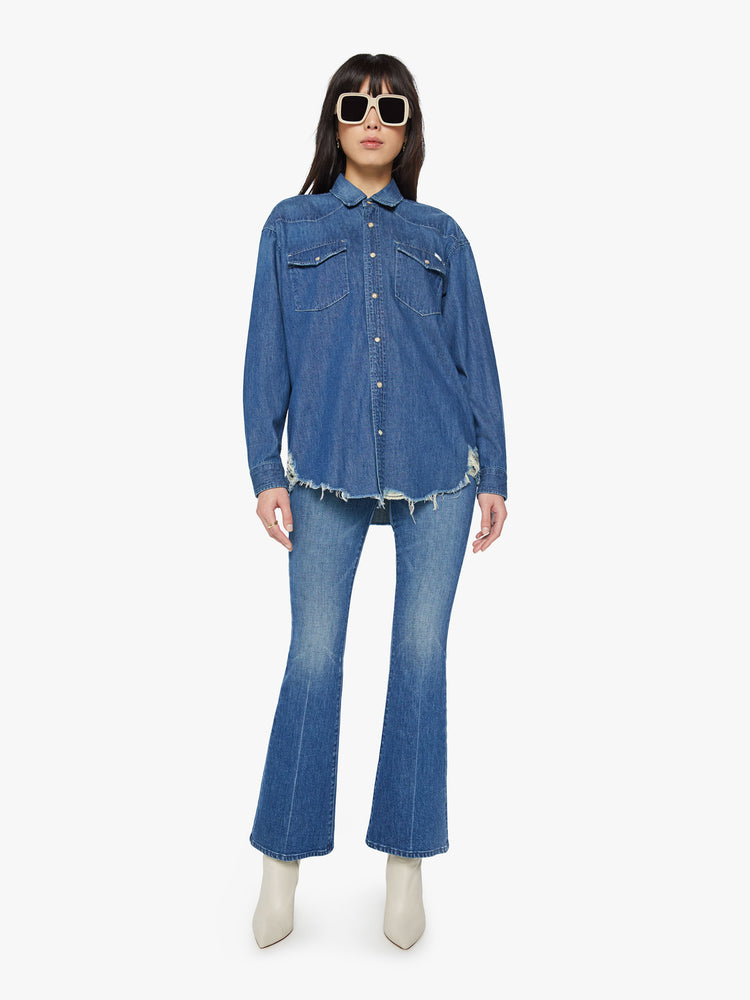 Full body view of a woman oversized western inspired denim button-up with front patch pockets and a raw, curved hem in a vintage blue wash.