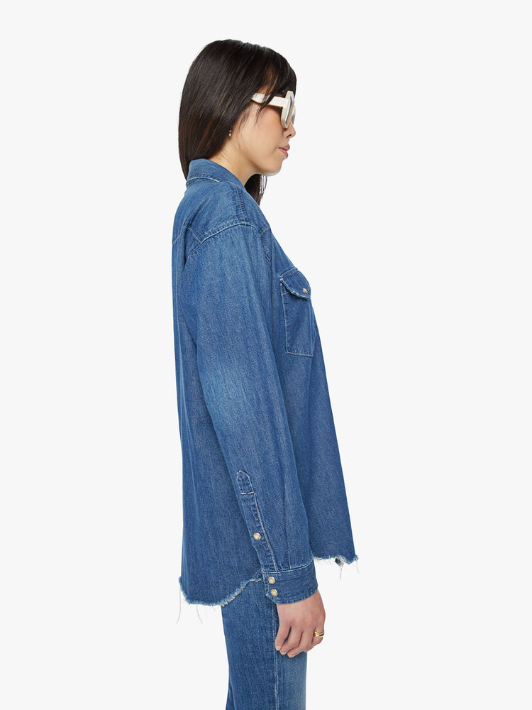 Side view of a woman oversized western inspired denim button-up with front patch pockets and a raw, curved hem in a vintage blue wash.