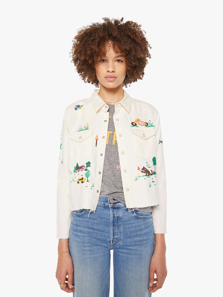 Front view of an off white with colorful embroidered scenes throughout denim button-up with front patch pockets and a cropped, raw hem.