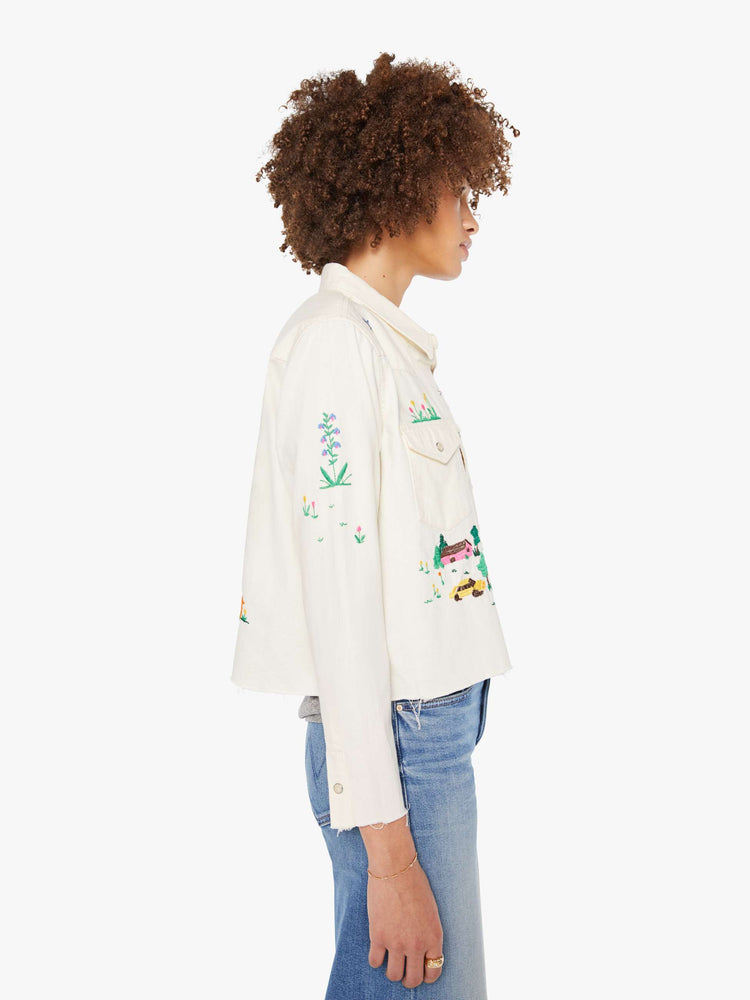 Side view of an off white with colorful embroidered scenes throughout denim button-up with front patch pockets and a cropped, raw hem.