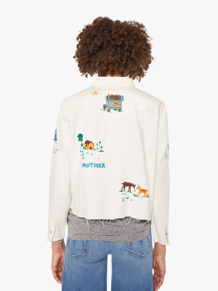 Back view of an off white with colorful embroidered scenes throughout denim button-up with front patch pockets and a cropped, raw hem.