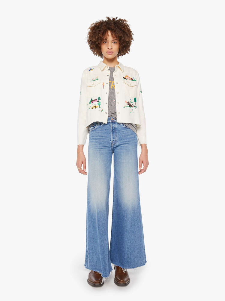 Full body view of an off white with colorful embroidered scenes throughout denim button-up with front patch pockets and a cropped, raw hem.