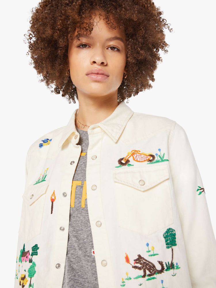 Close up view of an off white with colorful embroidered scenes throughout denim button-up with front patch pockets and a cropped, raw hem.