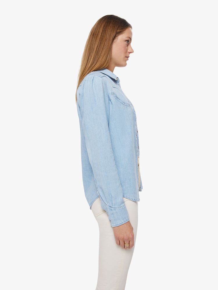 Side view of a woman denim button-up with rounded pockets, yoke detailing, slightly puffed sleeves and a curved hem in a light blue wash.