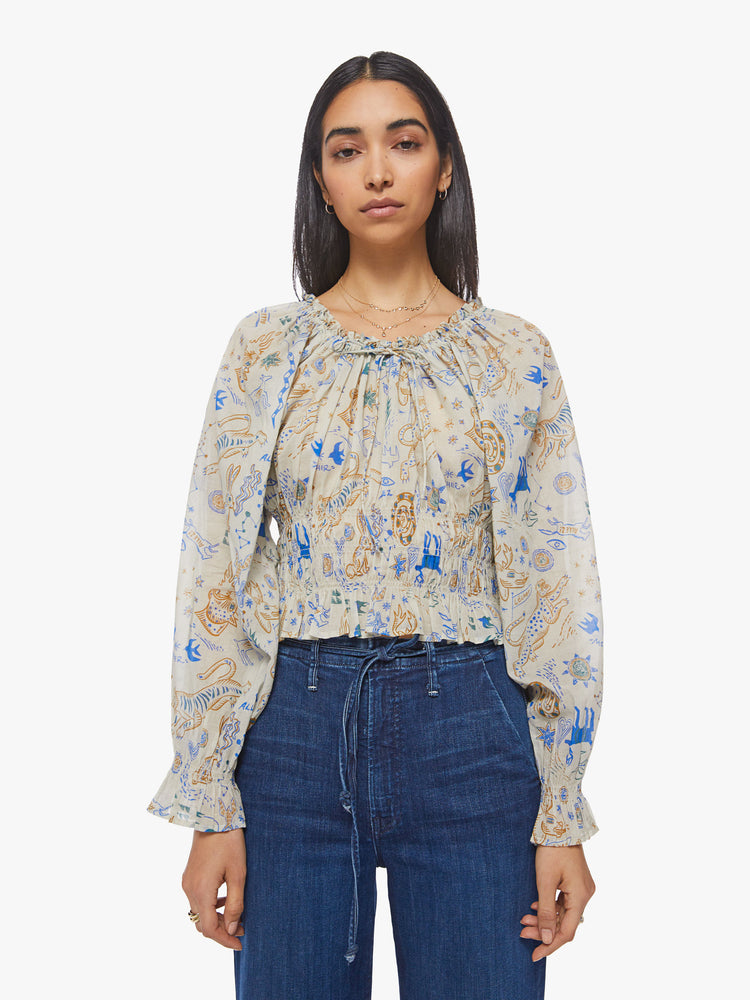 Front view of a woman creamy hue with animal doodles in blue and tan lightweight blouse with a gathered neckline that ties, long balloon sleeves, smocking at the waist and wrists and ruffled hems.