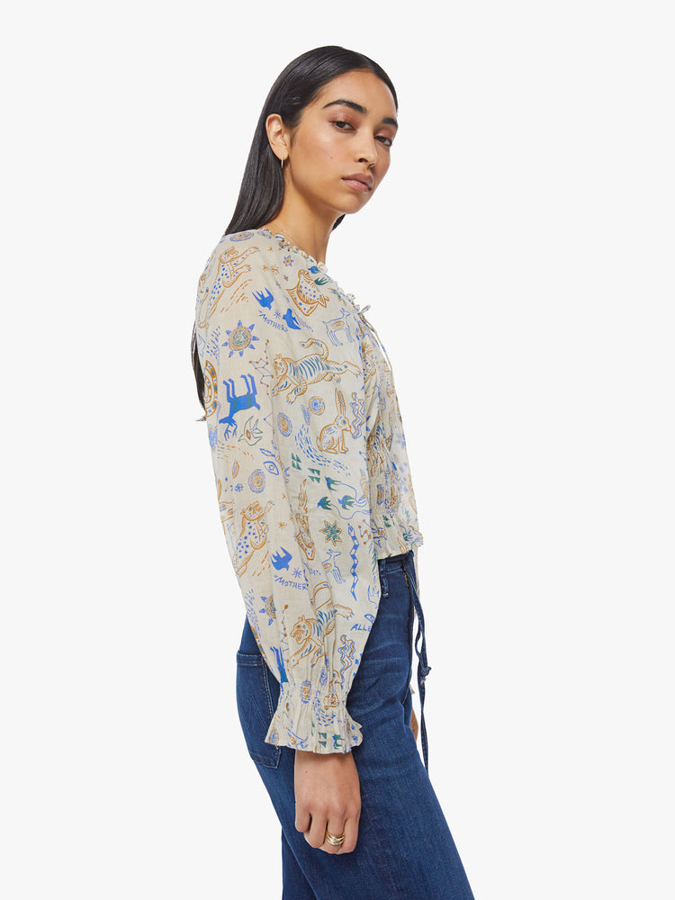 Side view of a woman creamy hue with animal doodles in blue and tan lightweight blouse with a gathered neckline that ties, long balloon sleeves, smocking at the waist and wrists and ruffled hems.