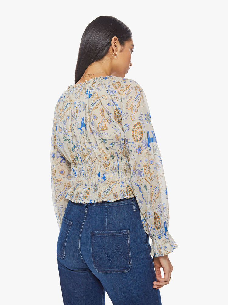 Back view of a woman creamy hue with animal doodles in blue and tan lightweight blouse with a gathered neckline that ties, long balloon sleeves, smocking at the waist and wrists and ruffled hems.