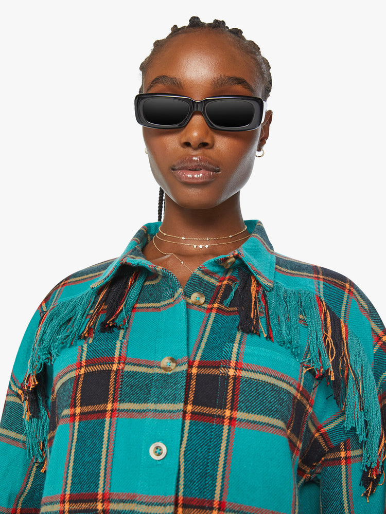 Close up view of a woman western inspired button-up with drop shoulders, a curved hem and fringe across the chest and back in a bright blue, red and yellow plaid paint.