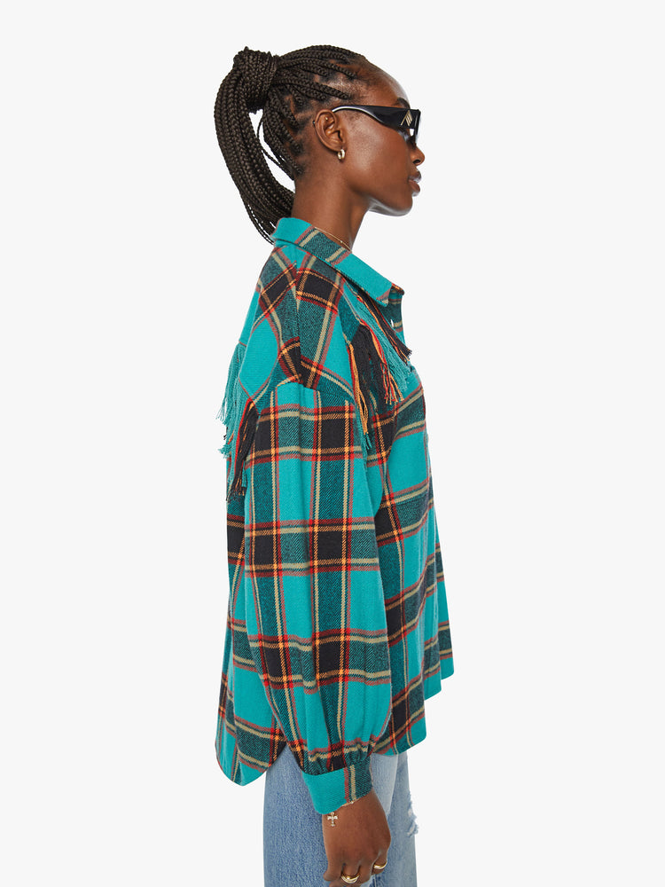 Side view of a woman western inspired button-up with drop shoulders, a curved hem and fringe across the chest and back in a bright blue, red and yellow plaid paint.