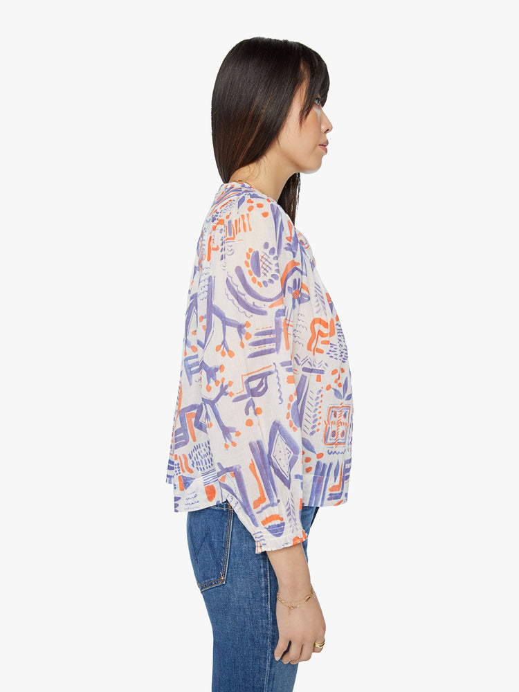 Side view of a woman flowy blouse with a V-neck, cropped balloon sleeves, buttons down the front and ruffles throughout for a loose fit in off white doodles in red and blue.