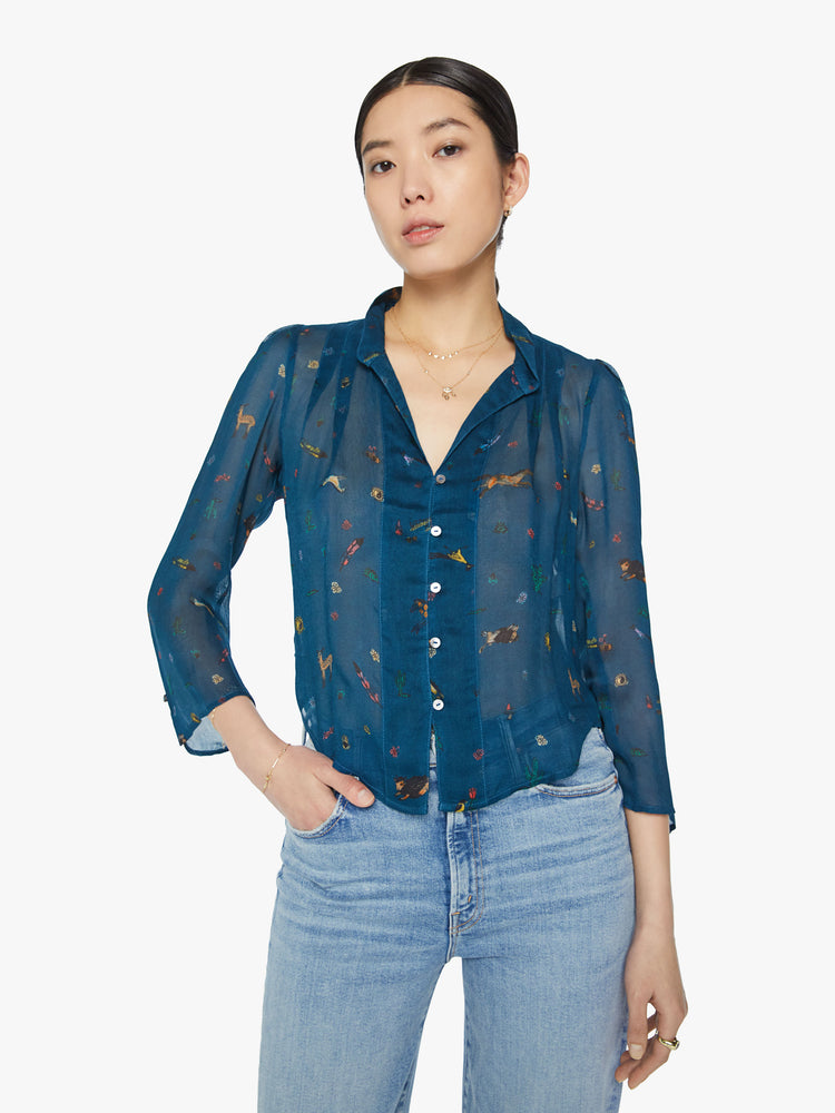 Front view of a woman in a semi sheer blue  button-up blouse with a shrunken collar, 3/4-length sleeves and a cropped curved hem with small horses, cacti, suns and more.