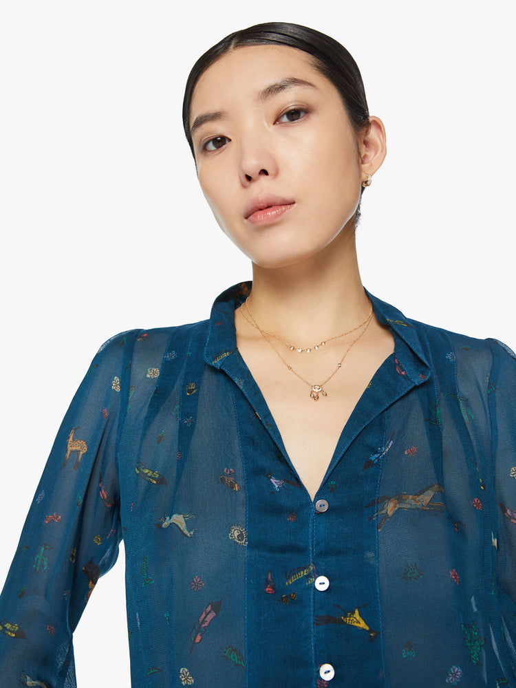Close up view of a woman in a semi sheer blue button-up blouse with a shrunken collar, 3/4-length sleeves and a cropped curved hem with small horses, cacti, suns and more.
