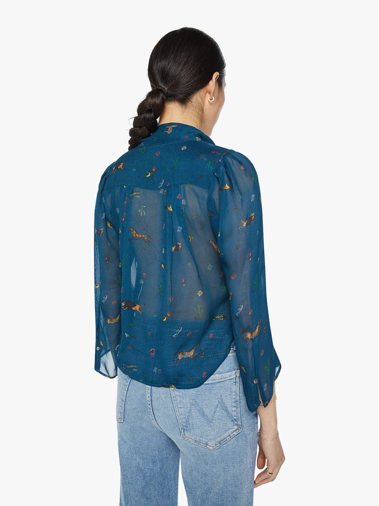 Back view of a woman in a semi sheer blue button-up blouse with a shrunken collar, 3/4-length sleeves and a cropped curved hem with small horses, cacti, suns and more.