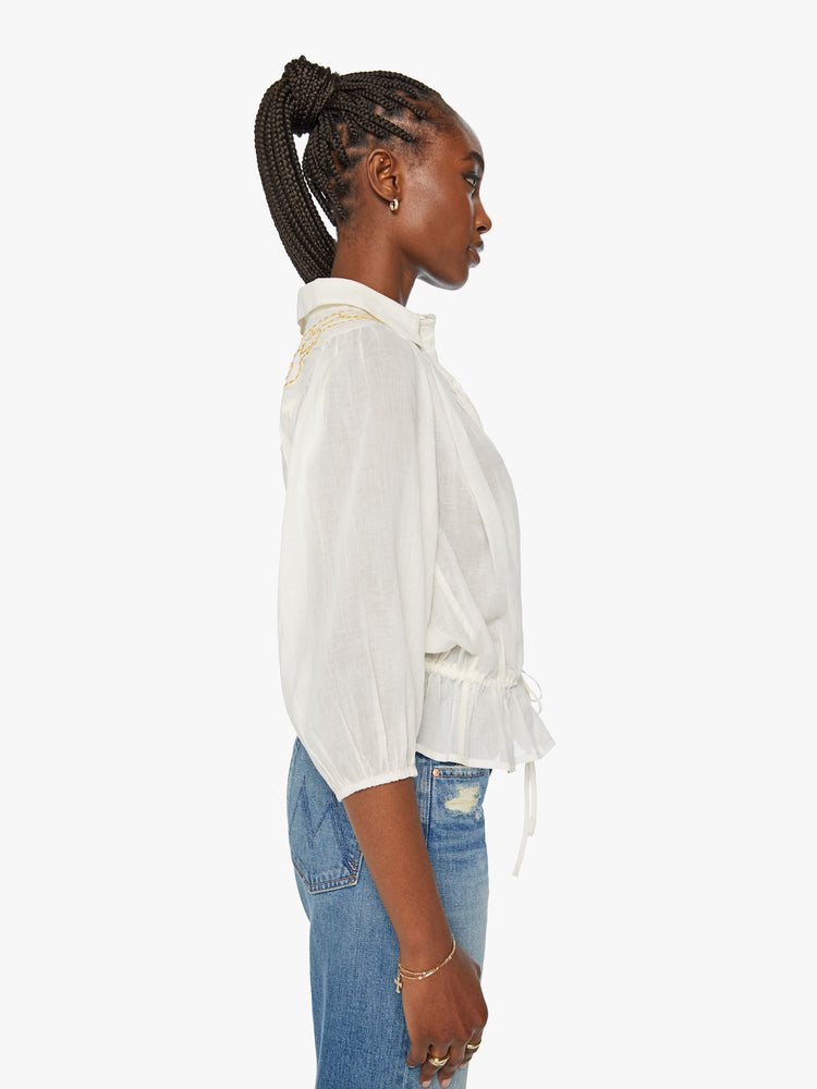 Side  view of a woman off white hue ollared blouse with a ruffled neckline, 3/4-length balloon sleeves, buttons down the front and a drawstring waist and orange applique dove on the back and shoulders.