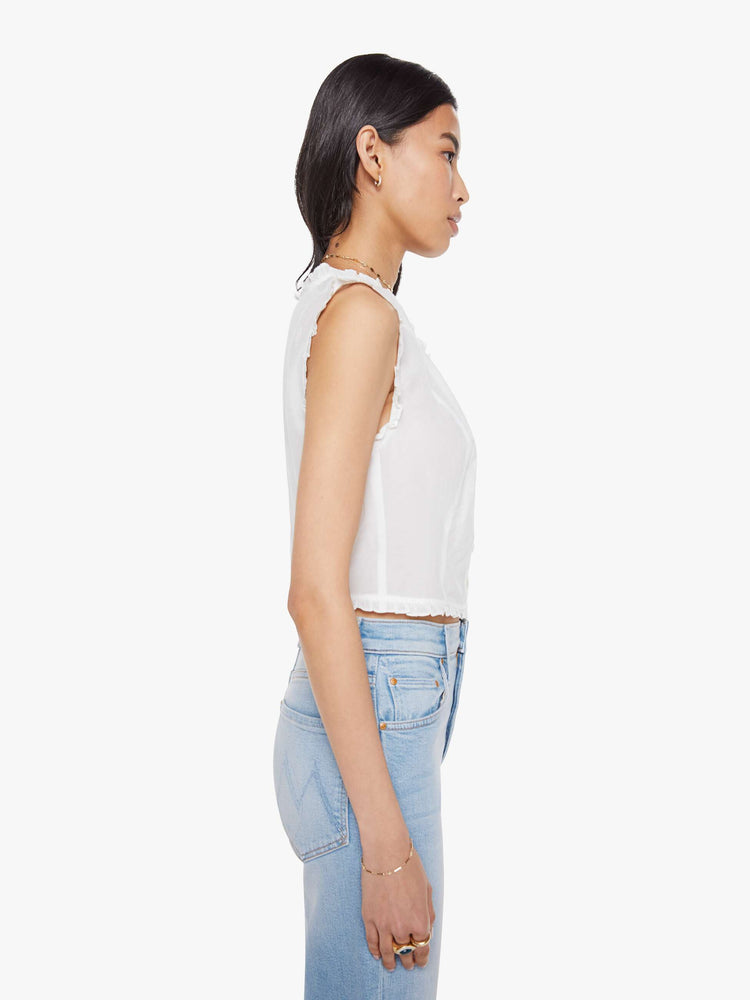 Side view of woman in a sheer white sleeveless scoop neck blouse with ruffled hems, seamed details, buttons down the front and a cropped fit.