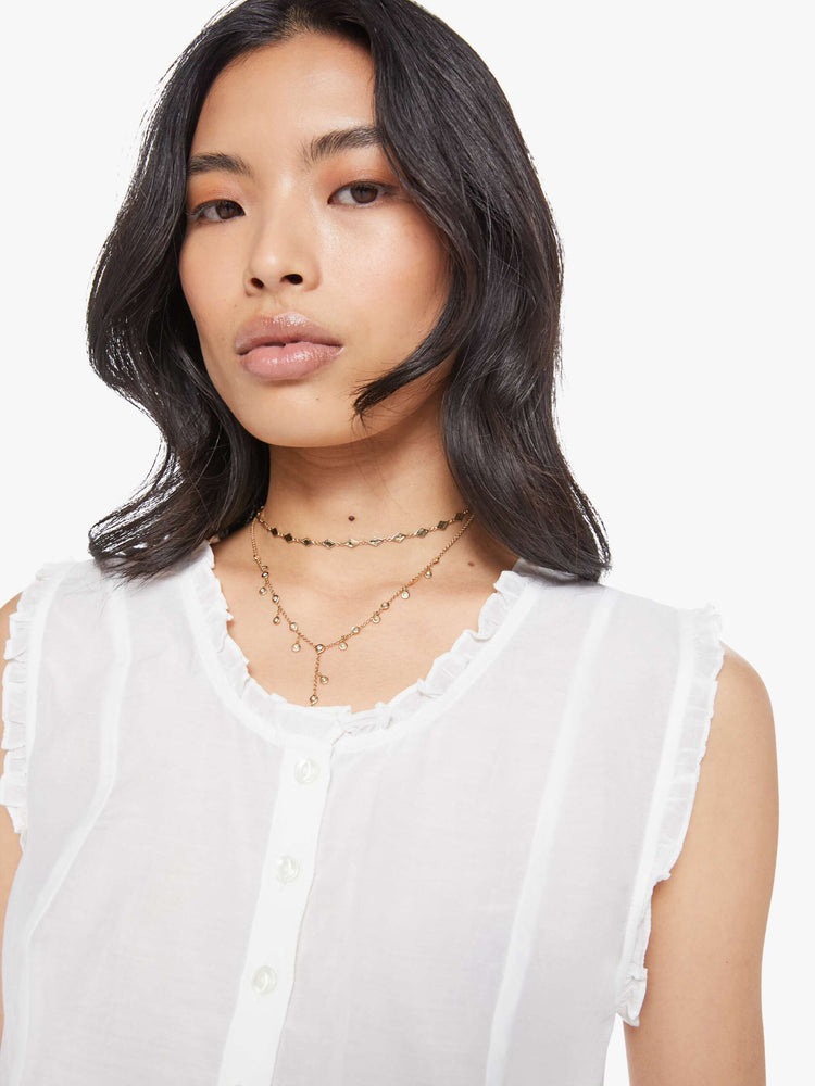 Close up view of woman in a sheer white sleeveless scoop neck blouse with ruffled hems, seamed details, buttons down the front and a cropped fit.