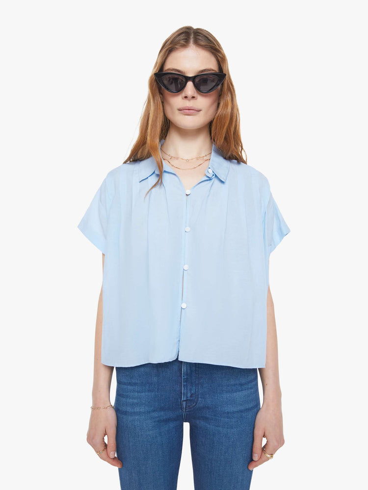 Front view of a woman in a baby blue boxy short-sleeved button-up with drop shoulders, pleats across the chest and back and a cropped hem.