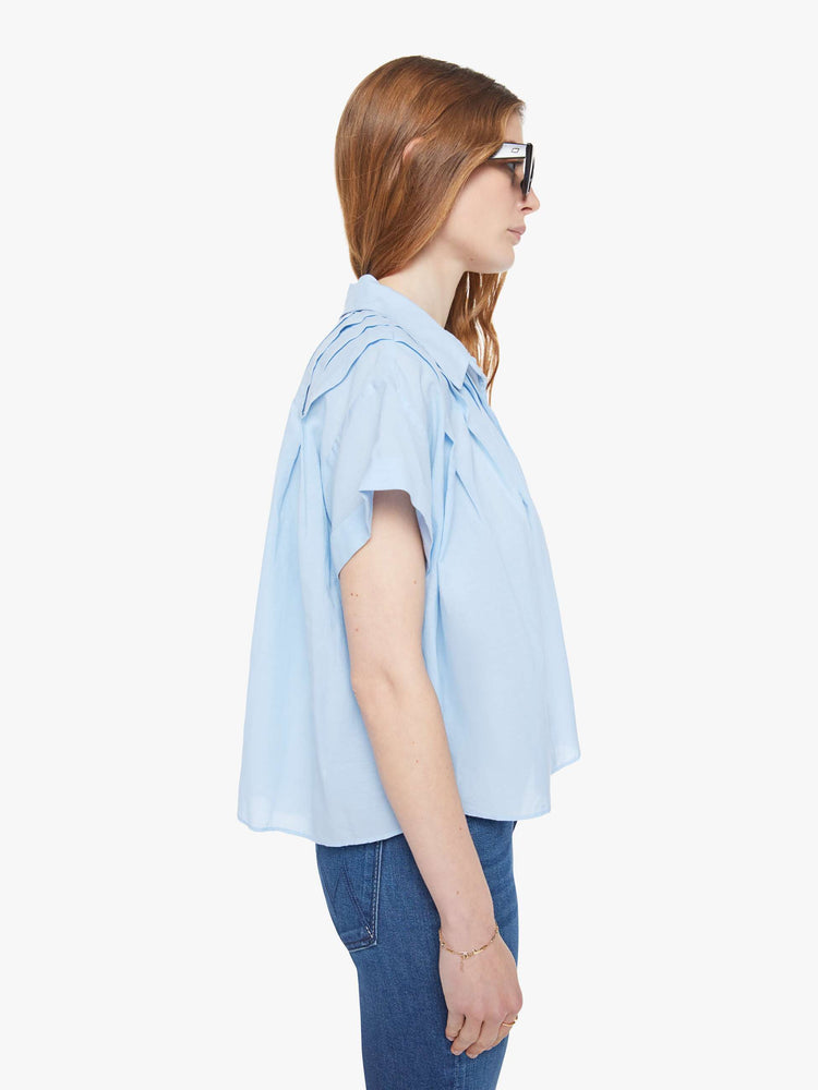 Side view of a woman in a baby blue boxy short-sleeved button-up with drop shoulders, pleats across the chest and back and a cropped hem.