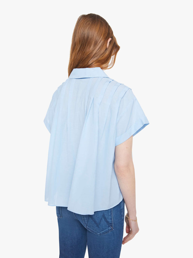 Back view of a woman in a baby blue boxy short-sleeved button-up with drop shoulders, pleats across the chest and back and a cropped hem.