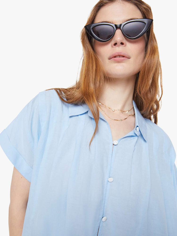Close up view of a woman in a baby blue boxy short-sleeved button-up with drop shoulders, pleats across the chest and back and a cropped hem.