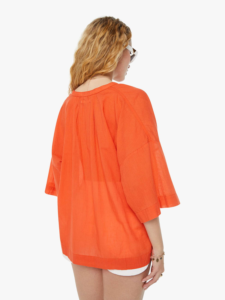 Back view of a woman in a sheer orange V-neck top with oversized patch pocket at the chest.