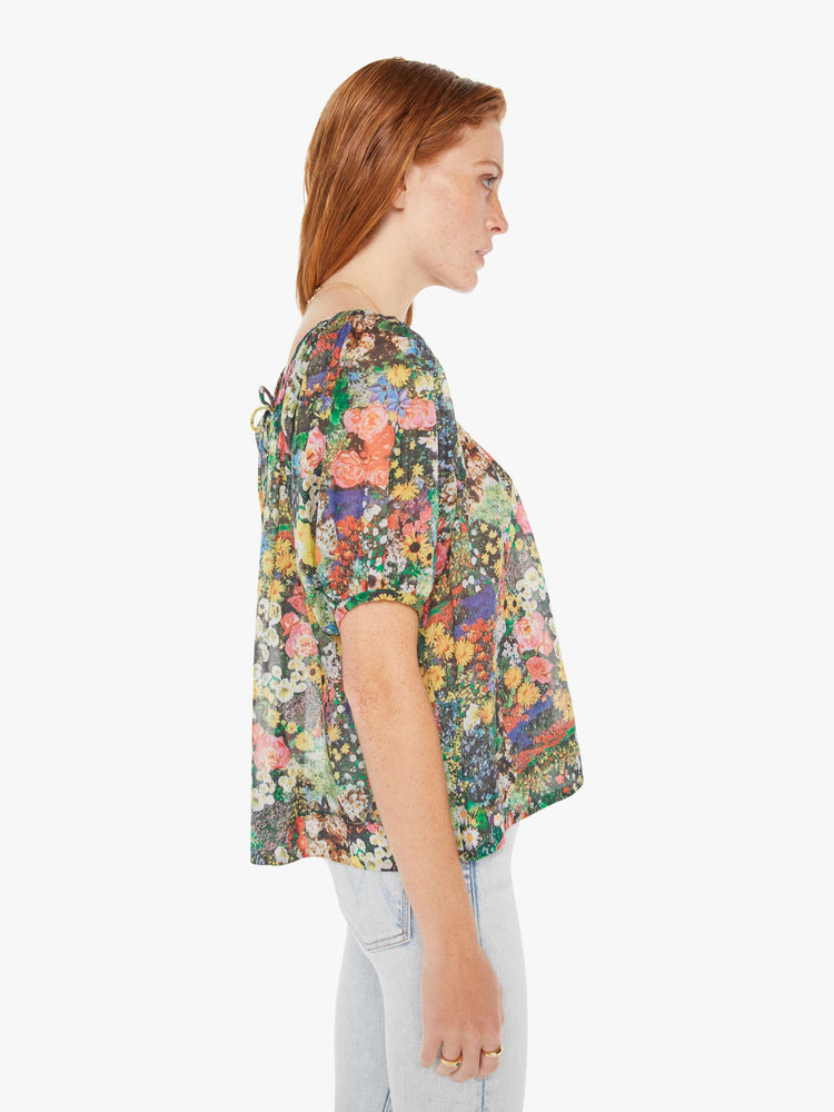 Side view of a woman wearing a sheer peasant blouse featuring a colorful garden print, paired with a light blue wash jean.
