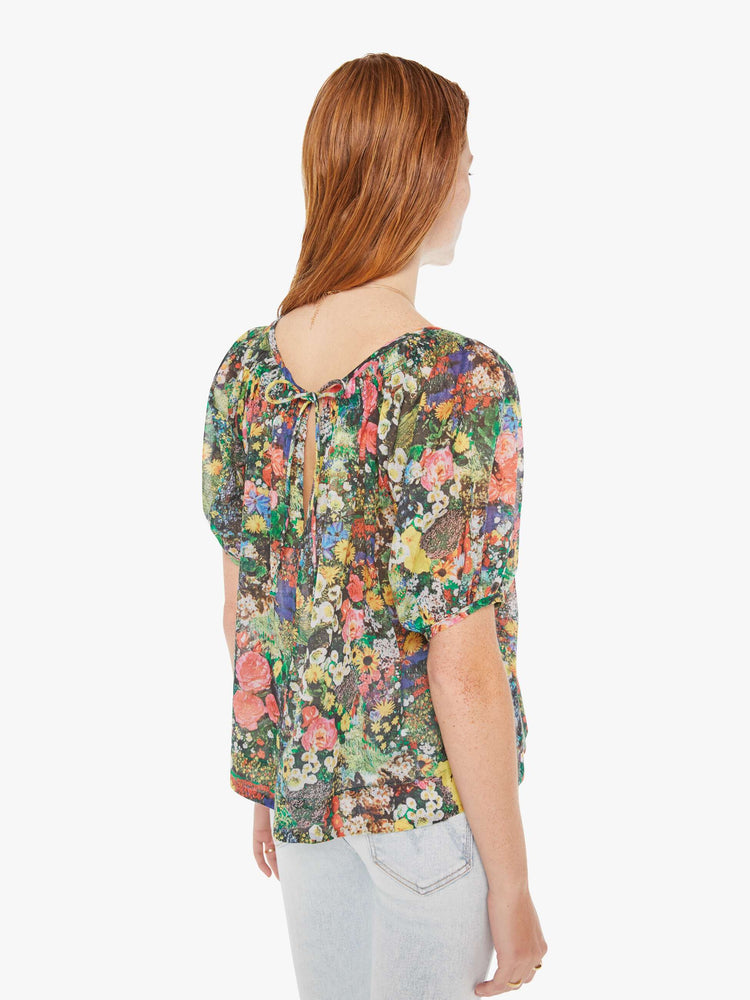 Back view of a woman wearing a sheer peasant blouse featuring a colorful garden print, paired with a light blue wash jean.