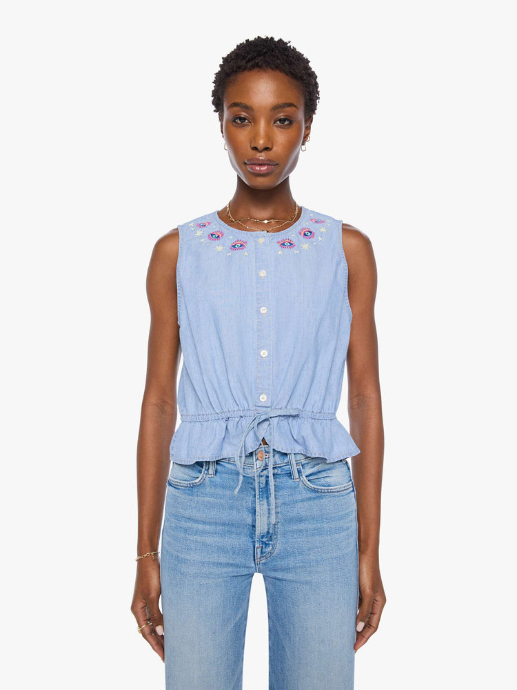 Front view of a woman in light blue with colorful embroidered eyes at the neckline denim tank with a crewneck, buttons down the front and a tie-waist detail.