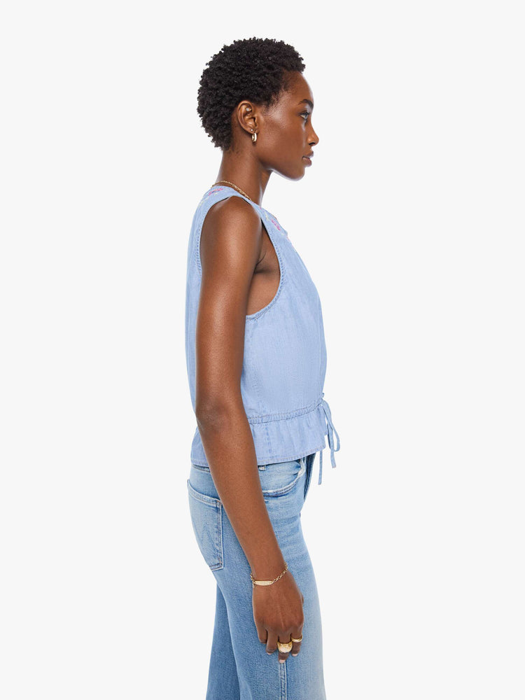 Side view of a woman in light blue with colorful embroidered eyes at the neckline denim tank with a crewneck, buttons down the front and a tie-waist detail.