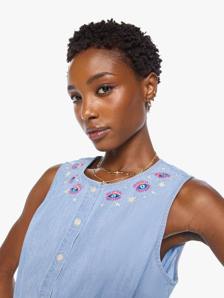 Close up view of a woman in light blue with colorful embroidered eyes at the neckline denim tank with a crewneck, buttons down the front and a tie-waist detail.