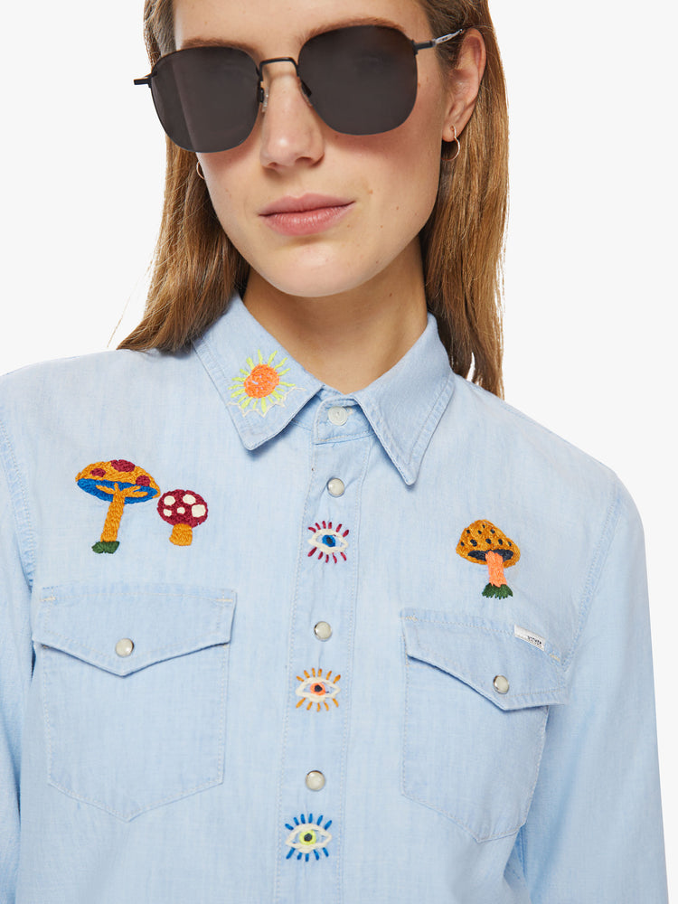 Close up view of a western denim button up with front patch pockets, a curved hem, and snap closures down the front and at the wrists in a light blue with embroidery on chest and shoulders.