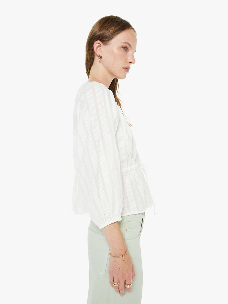 Side view of a woman in a white scoopneck blouse with 3/4-length balloon sleeves, ruffles throughout, tied details at the neck and waist and a slightly cropped hem.