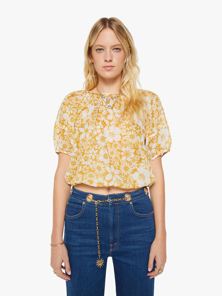Front view of a woman in an off-white with a golden yellow floral print blouse with a gathered crewneck, short balloon sleeves, a cropped elastic hem and an open back that ties at the neck.