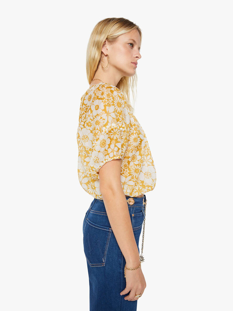 Side view of a woman in an off-white with a golden yellow floral print blouse with a gathered crewneck, short balloon sleeves, a cropped elastic hem and an open back that ties at the neck.