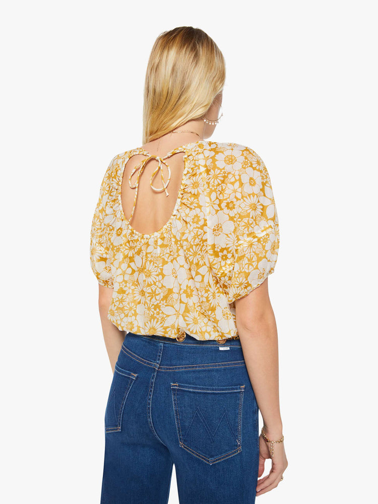 Back view of a woman in an off-white with a golden yellow floral print blouse with a gathered crewneck, short balloon sleeves, a cropped elastic hem and an open back that ties at the neck.