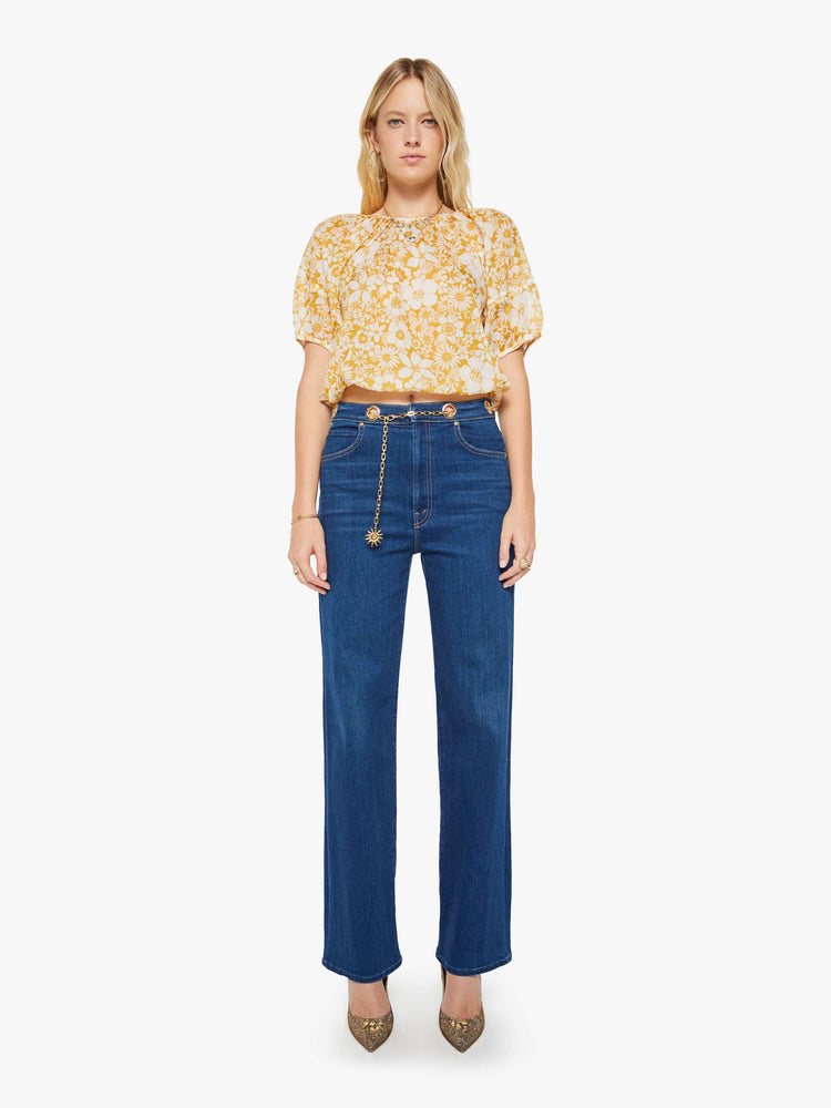 Full body view of a woman in an off-white with a golden yellow floral print blouse with a gathered crewneck, short balloon sleeves, a cropped elastic hem and an open back that ties at the neck.