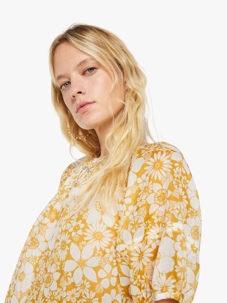Close up view of a woman in an off-white with a golden yellow floral print blouse with a gathered crewneck, short balloon sleeves, a cropped elastic hem and an open back that ties at the neck.