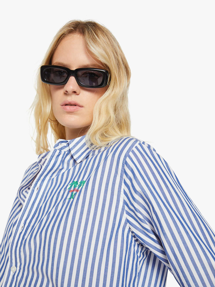 Close up view of a woman in a blue and white stripes button-up shirt with a curved hem and MOTHER's name embroidered with two small palm trees on the chest.