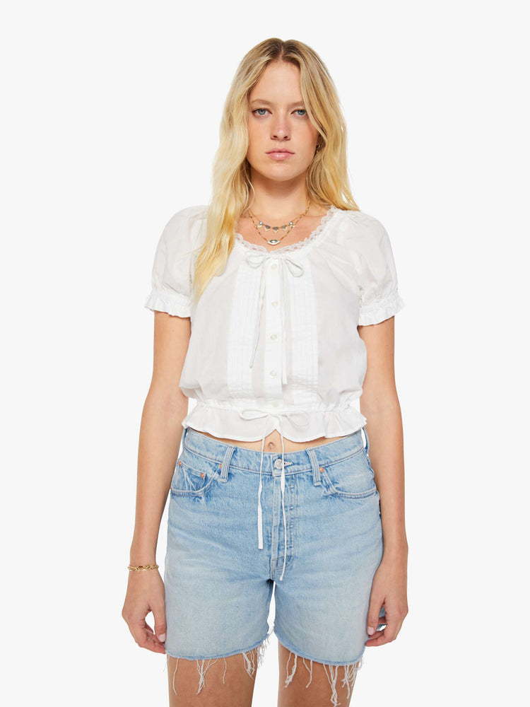 Front view of a woman in a white cropped blouse with a lace-trimmed scoop neck, short balloon sleeves, buttons down the front, a tied neck and waistband and ruffles at the hems.