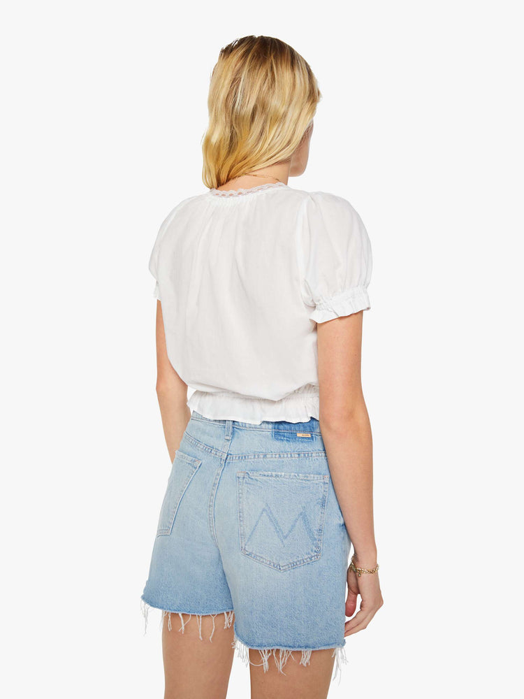 Back view of a woman in a white cropped blouse with a lace-trimmed scoop neck, short balloon sleeves, buttons down the front, a tied neck and waistband and ruffles at the hems.
