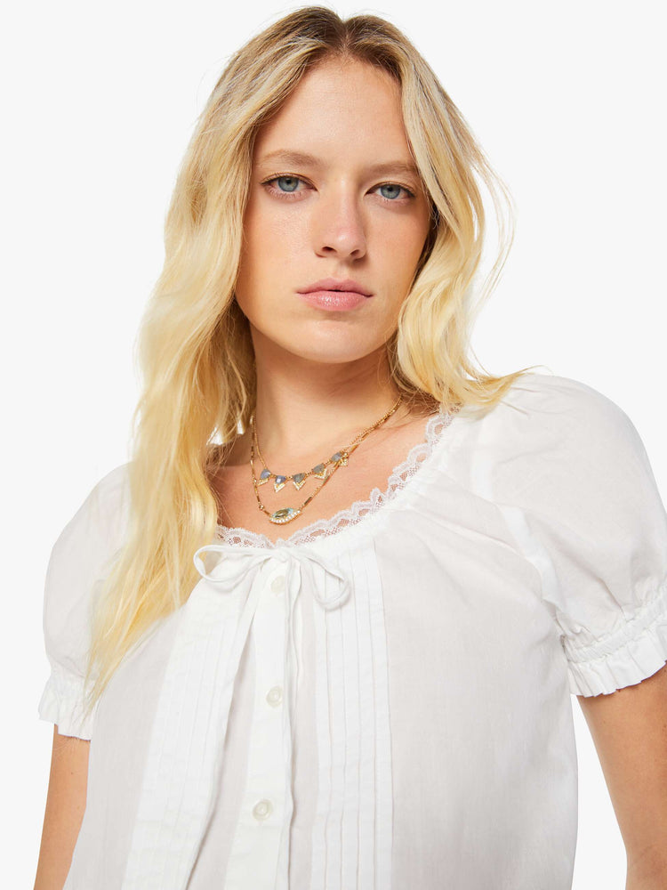 Close up view of a woman in a white cropped blouse with a lace-trimmed scoop neck, short balloon sleeves, buttons down the front, a tied neck and waistband and ruffles at the hems.