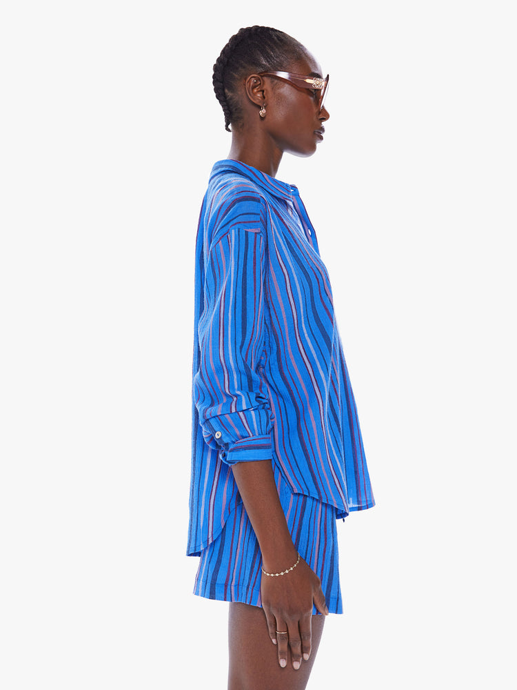 Side view of a women blue stripe pattern button down top with drop shoulders, a curved hem and a slightly oversized fit.