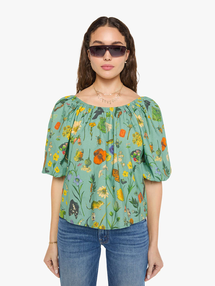 Front view of a woman in a teal-green with oversized printed flowers, gems, and creatures throughout flowy blouse.